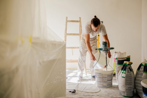 A constructor is standing in a house in a renovation process and adding water to a plaster mixture and preparing for skim coating. A worker is mixing materials in a bucket and preparing for plastering
